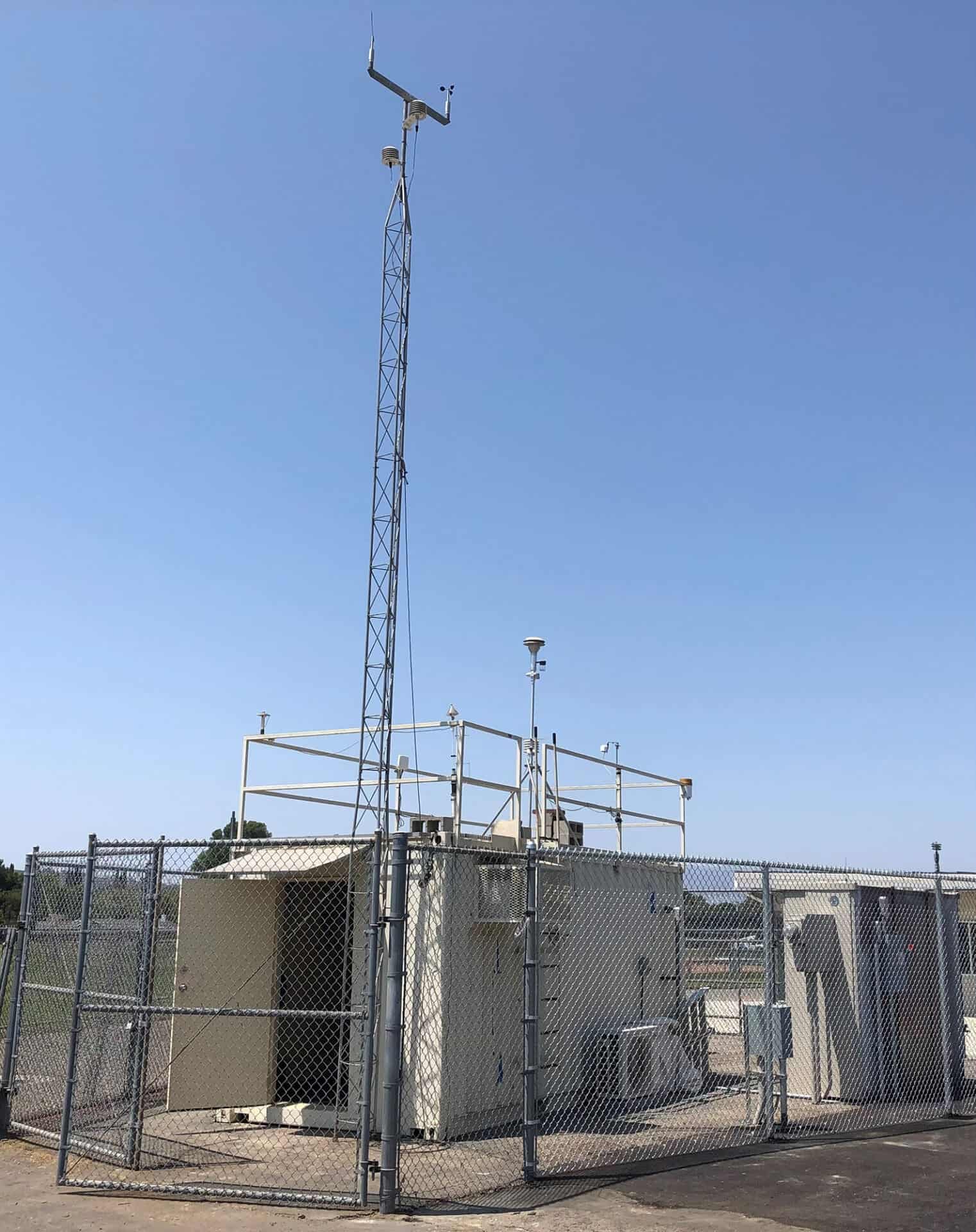 A fenced weather station with instruments on a rooftop and a tall antenna against a clear blue sky.