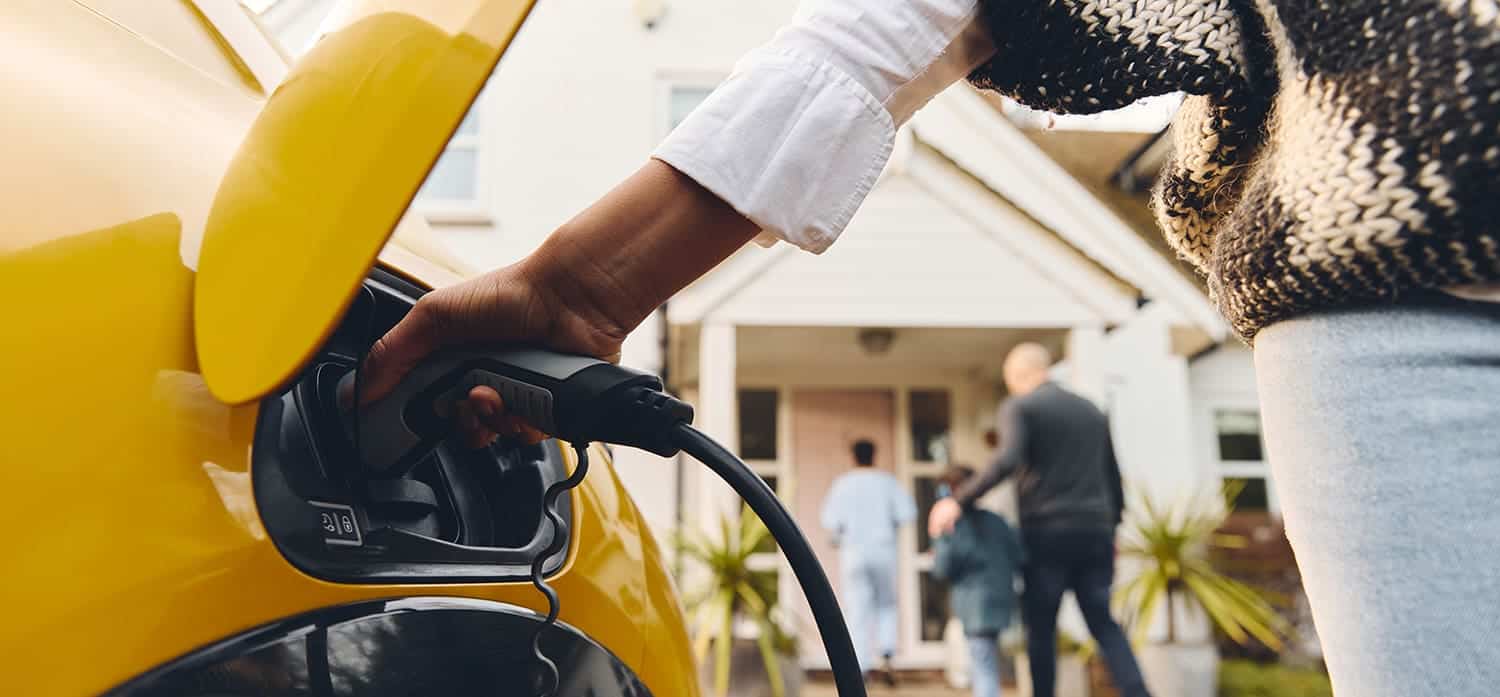 Person charging a yellow electric vehicle outside a house. Family in the background entering the home.