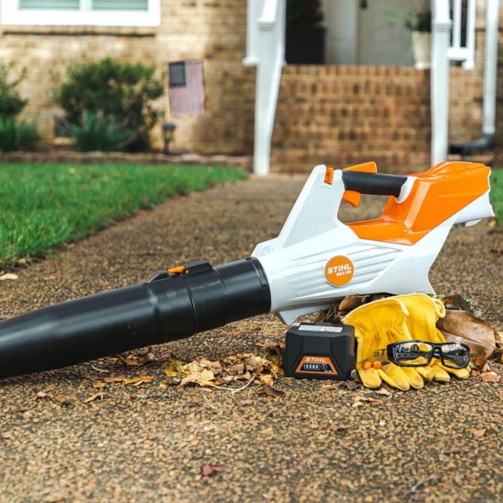 Leaf blower with accessories, including a battery, gloves, and safety glasses, on a sidewalk with scattered leaves, leading to a house entrance.