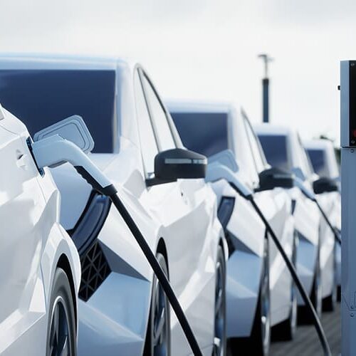 Row of electric cars charging at stations, with connected cables and green lights indicating active charge.