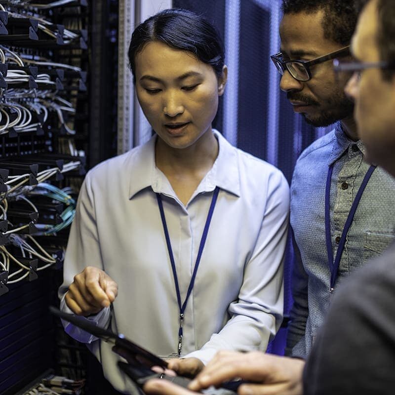 Three people in a server room discussing data on a digital tablet, surrounded by networking equipment and cables.