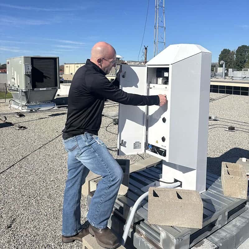 Man in black shirt opens door of metal box on a rooftop and adjusts a knob on it.
