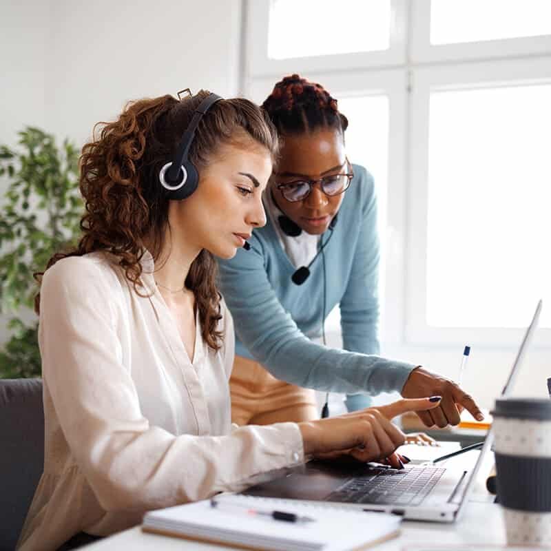 Two women wearing headsets work together at a laptop in a bright office. One woman is seated, the other stands beside her, both focused on the screen.